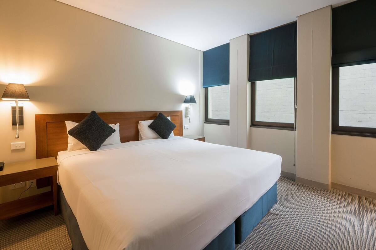 Quarto de hotel com uma cama grande, duas mesas de cabeceira, uma luminária na parede e uma janela com vista.