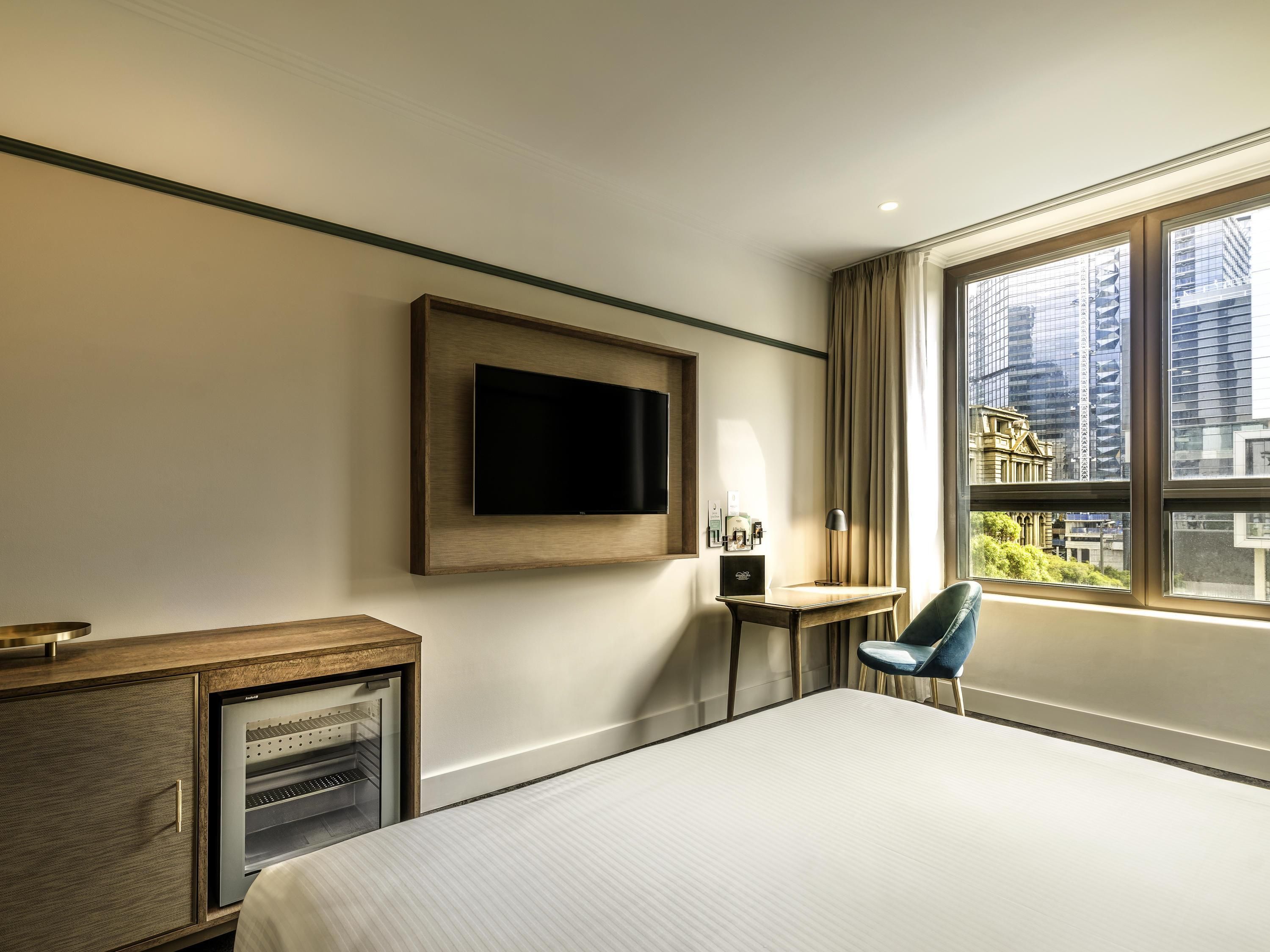 A hotel room with a bed, a TV mounted on the wall, a small table with a lamp, and a view of the city through large windows.