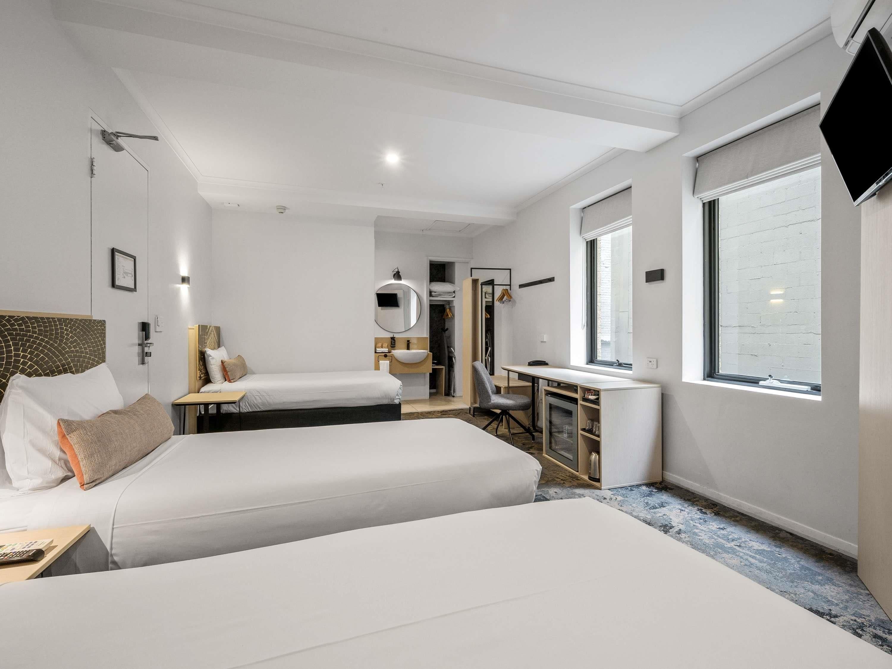 Ein Hotelzimmer mit zwei Betten, einem Schreibtisch, einem Stuhl, einem runden Spiegel, einem Fenster und einem Fernseher.