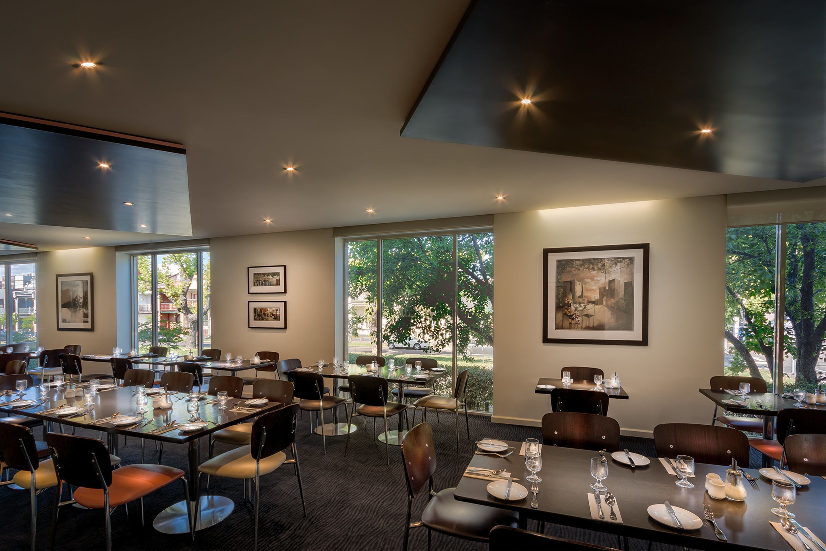 A modern dining area with multiple tables and chairs, large windows with a view of trees, and framed artwork on the walls.