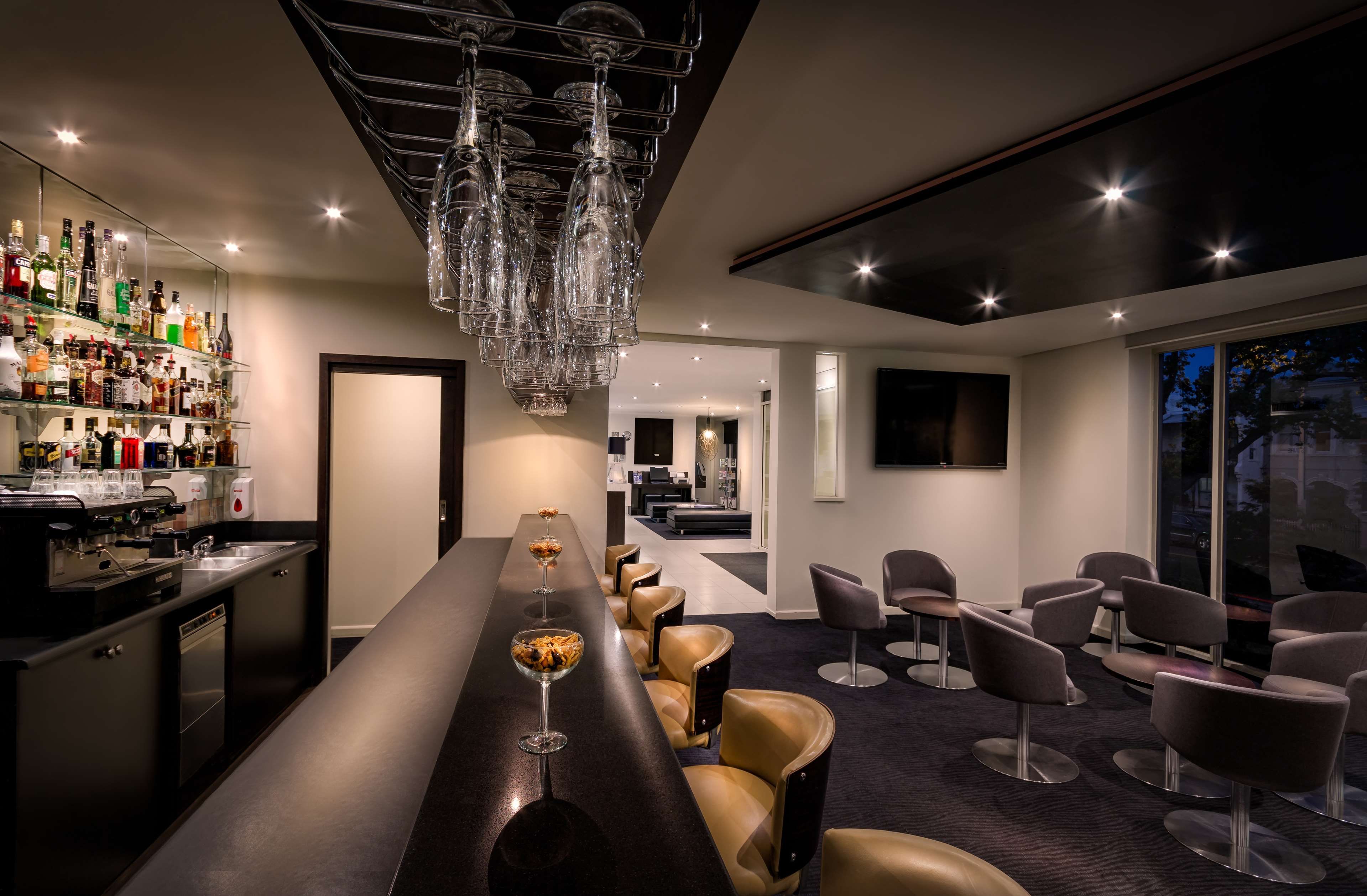 A bar area with a long counter, multiple glasses, and a variety of bottles. There are seating areas with chairs and a television mounted on the wall.