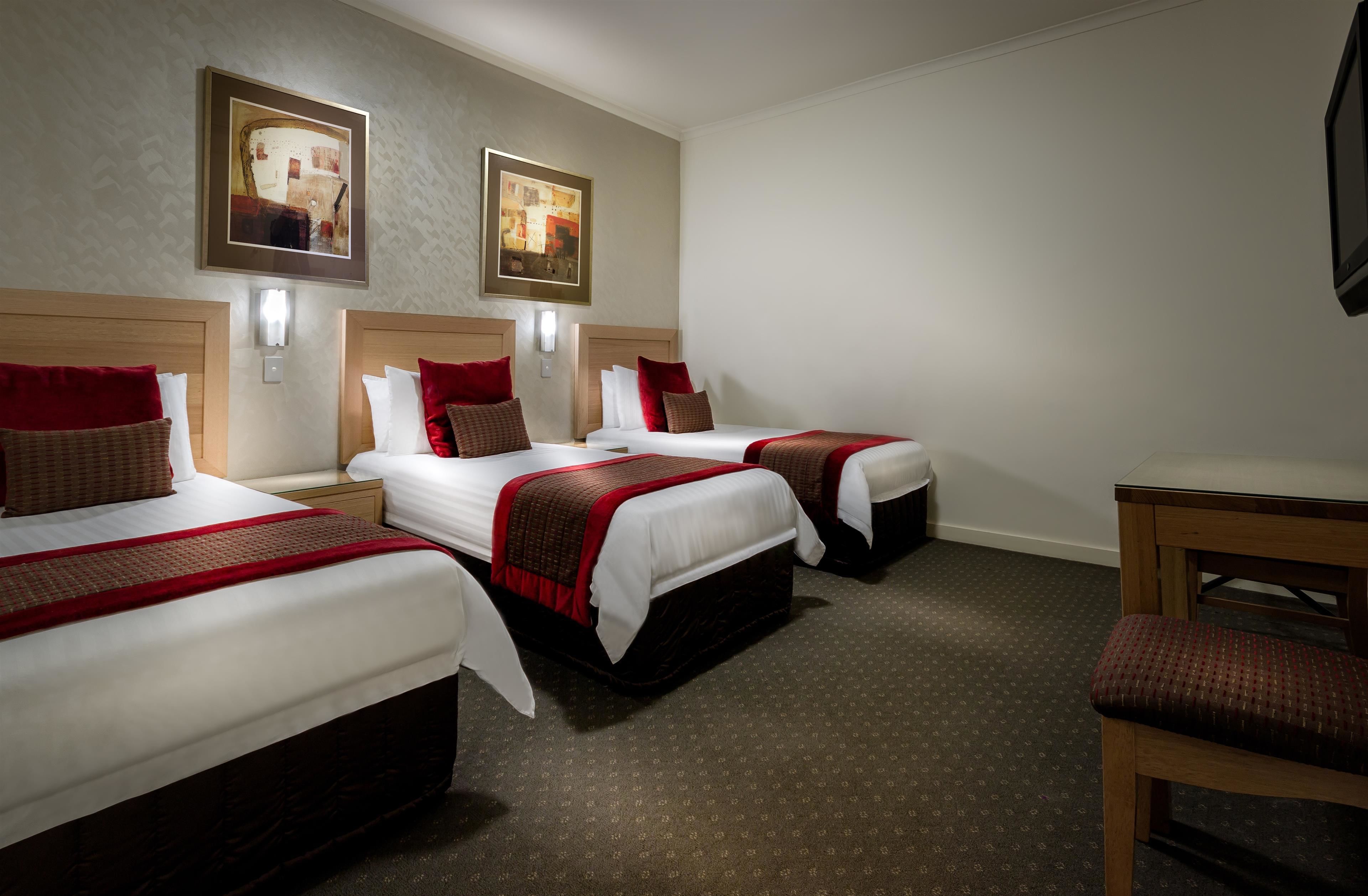 A hotel room with three beds, each with a red and white bedspread and matching pillows. There are two wall-mounted light fixtures and two framed pictures on the wall.