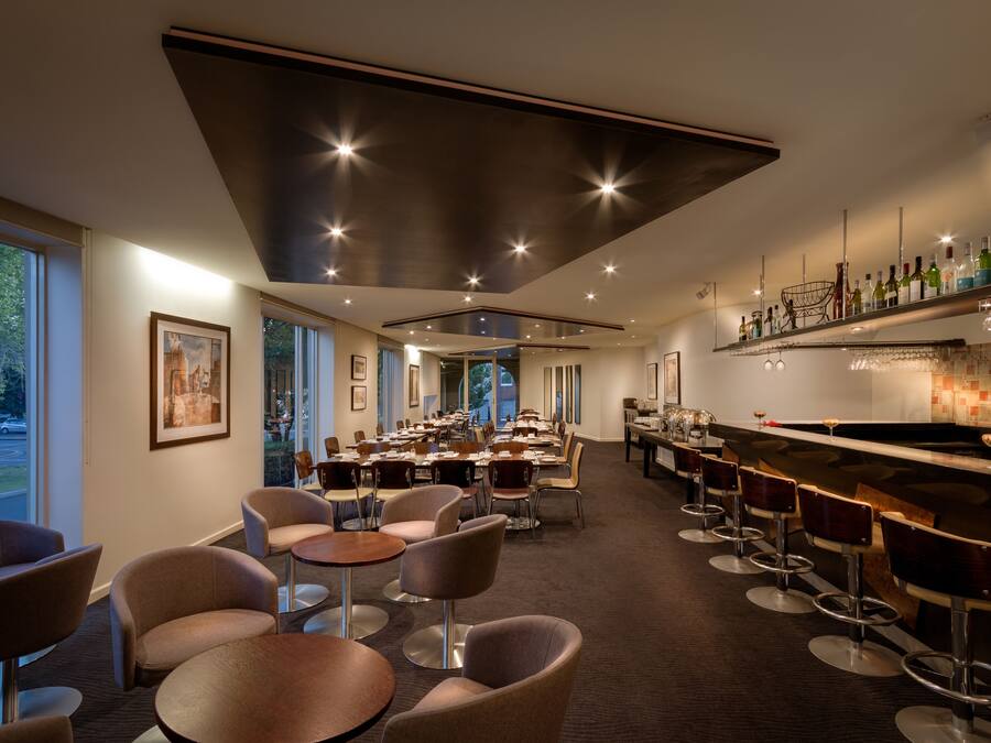 Un comedor moderno con mesas redondas y taburetes de bar, ventanas amplias y una barra con estantes llenos de botellas.