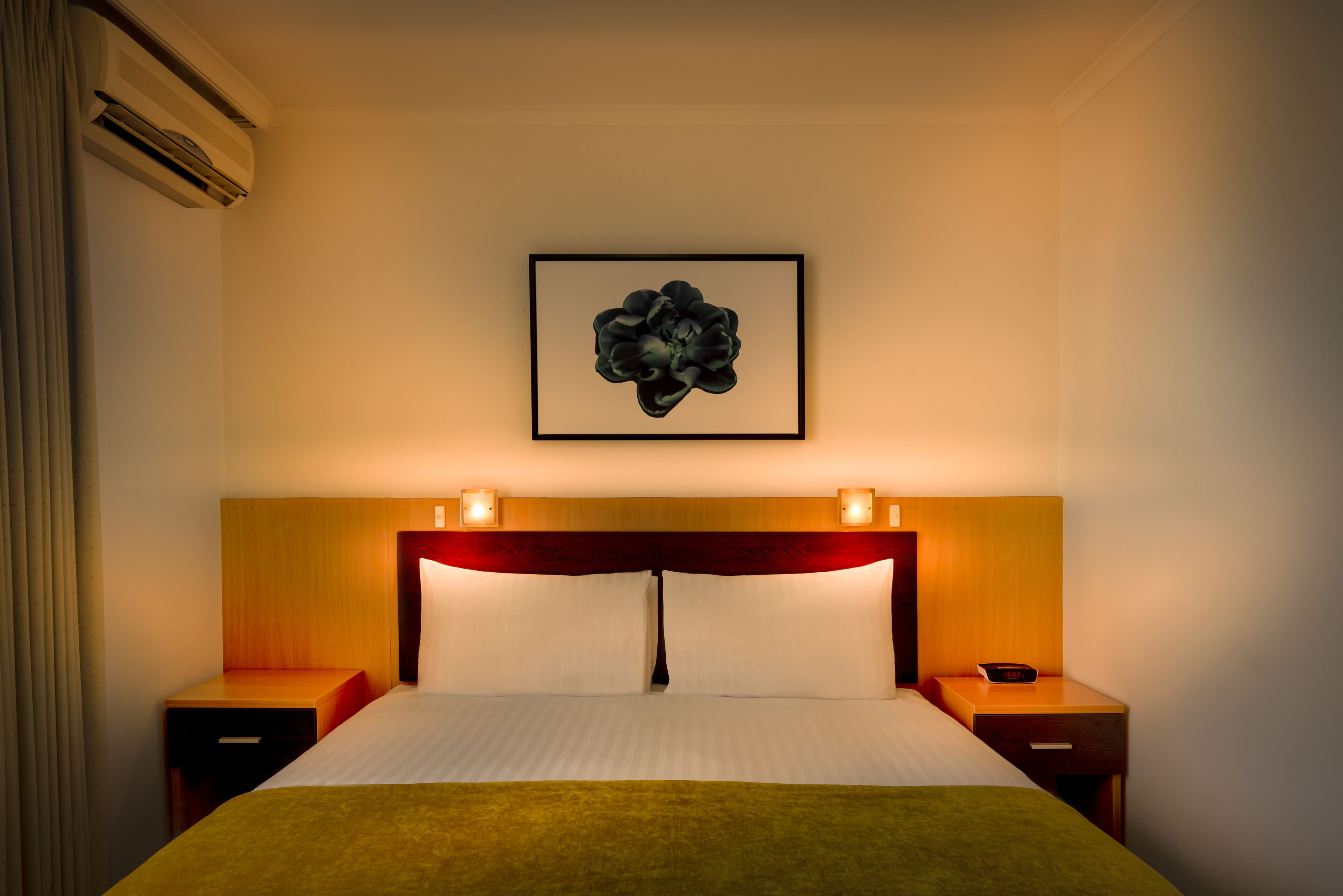A hotel room with a bed, two bedside tables, a wall-mounted air conditioner, and a framed artwork above the bed.