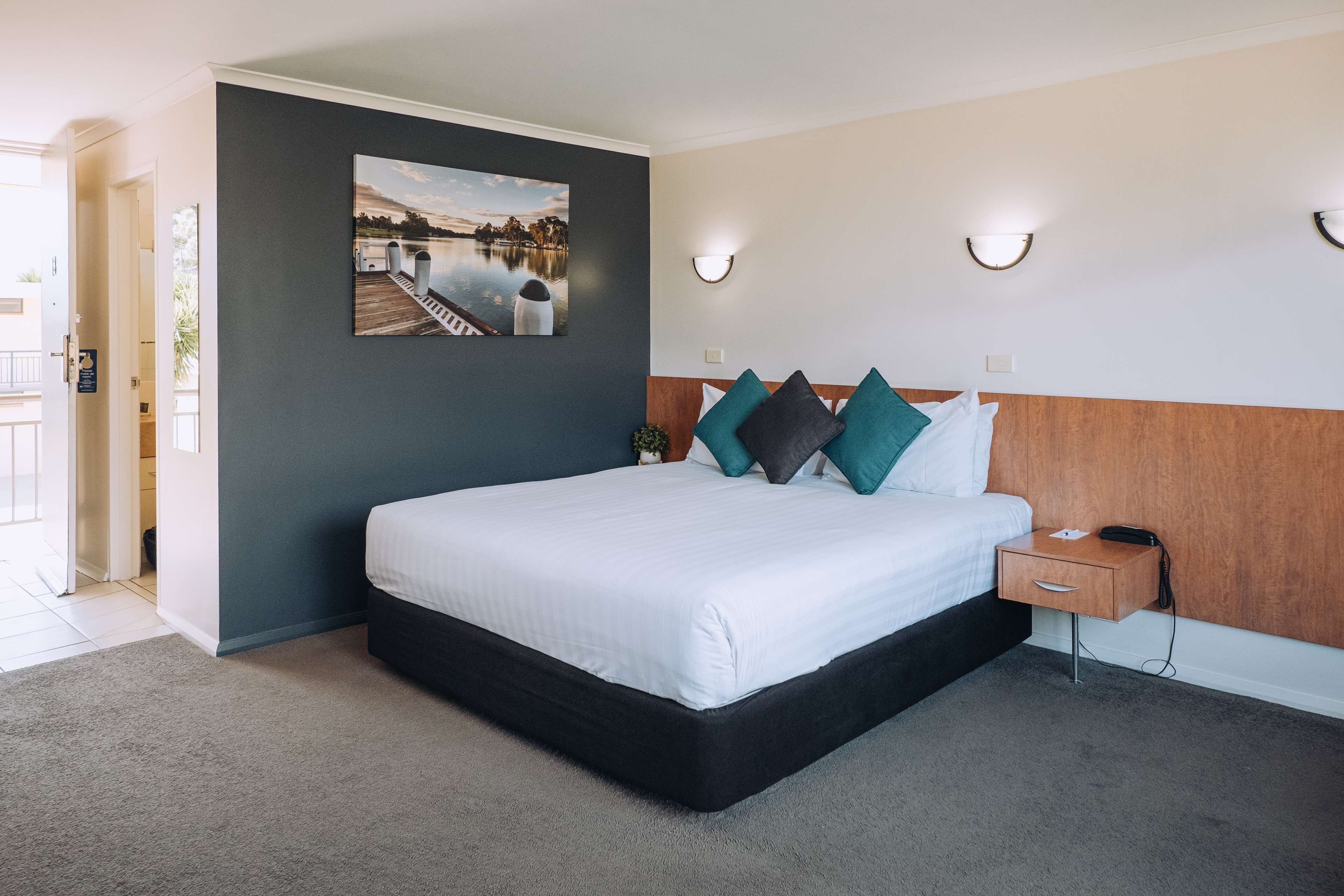 Habitación de hotel ordenada con una cama grande, una mesita de noche y una obra de arte en la pared que representa una escena junto al lago.
