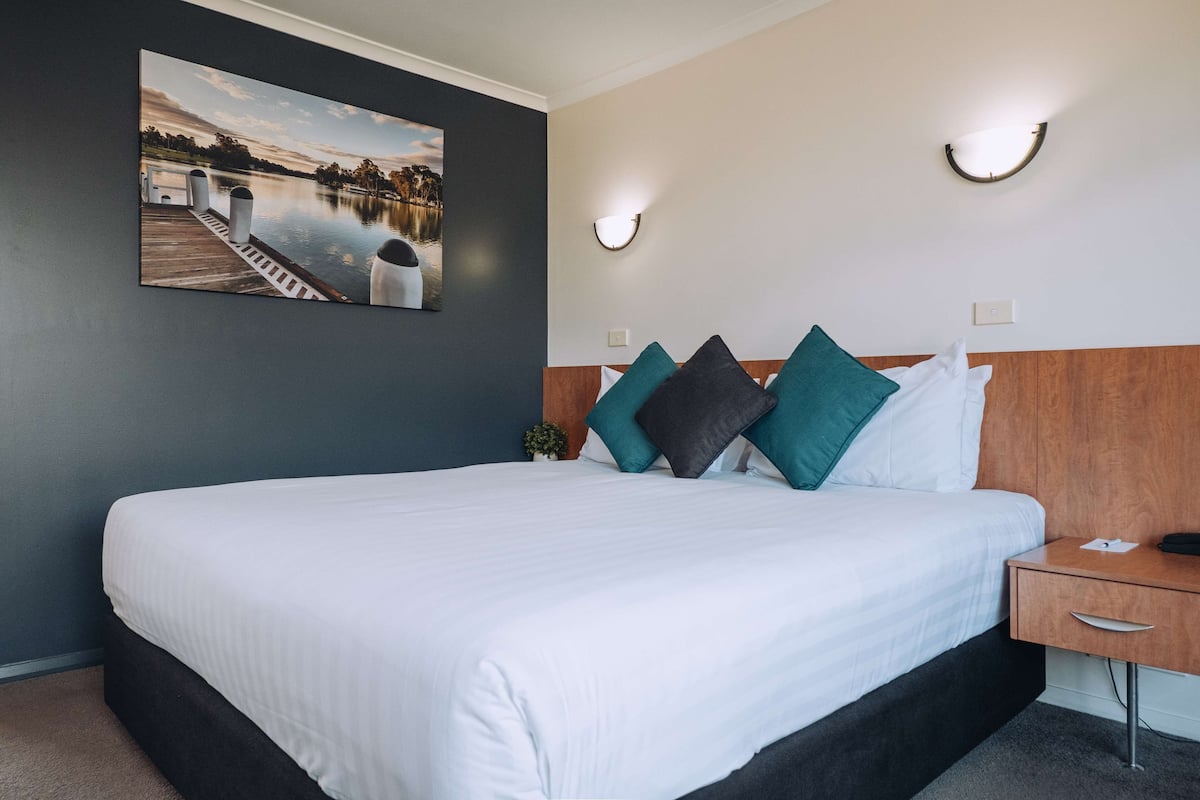 A neatly made bed with white bedding, a wooden headboard, and teal and black pillows. There is a small bedside table with a lamp and a phone. A framed picture of a lakeside scene hangs on the wall above the bed.