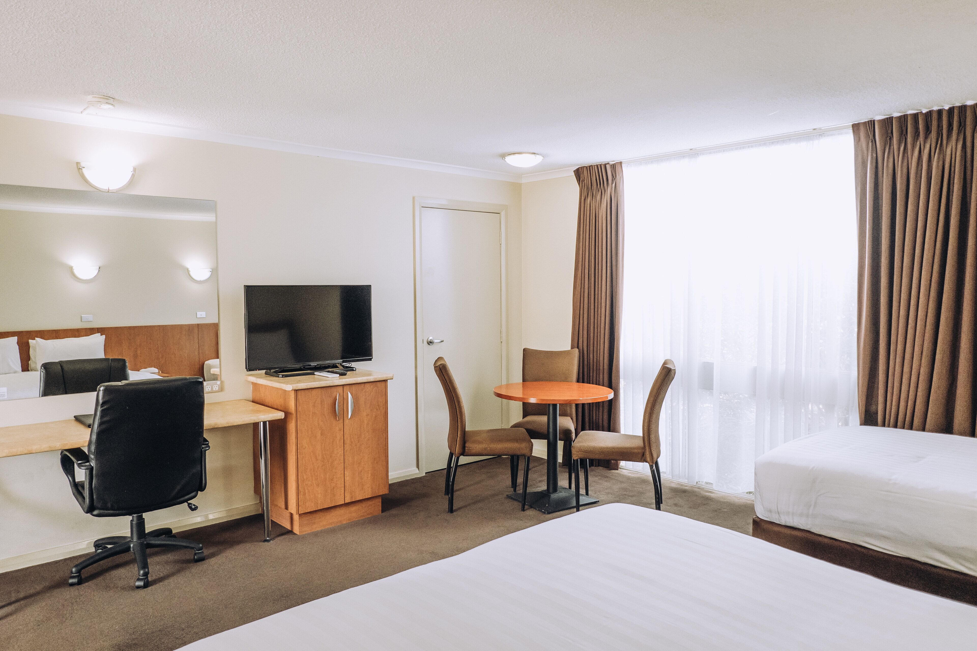 Ett hotellrum med en säng, ett skrivbord, en stol, en tv och ett fönster med gardiner.