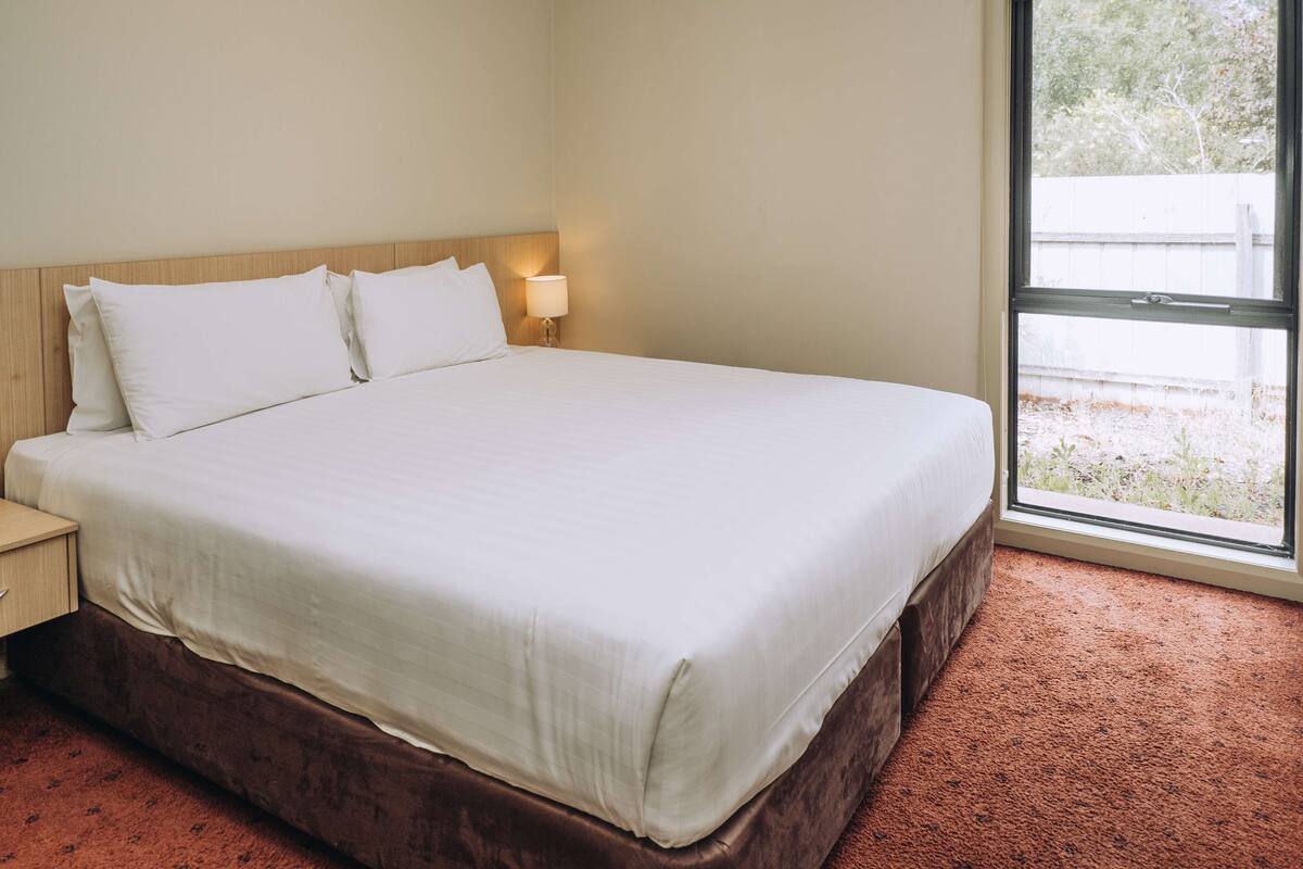 A neatly made bed with white linens in a room with a large window and a wooden headboard.