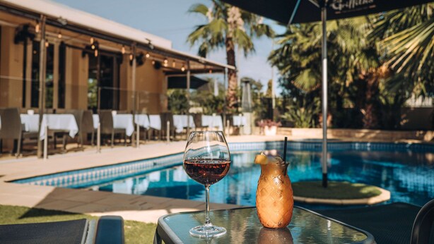 Un verre de vin rouge et une boisson tropicale sur une table placée au bord d’une piscine, avec un palmier et un bâtiment en arrière-plan.