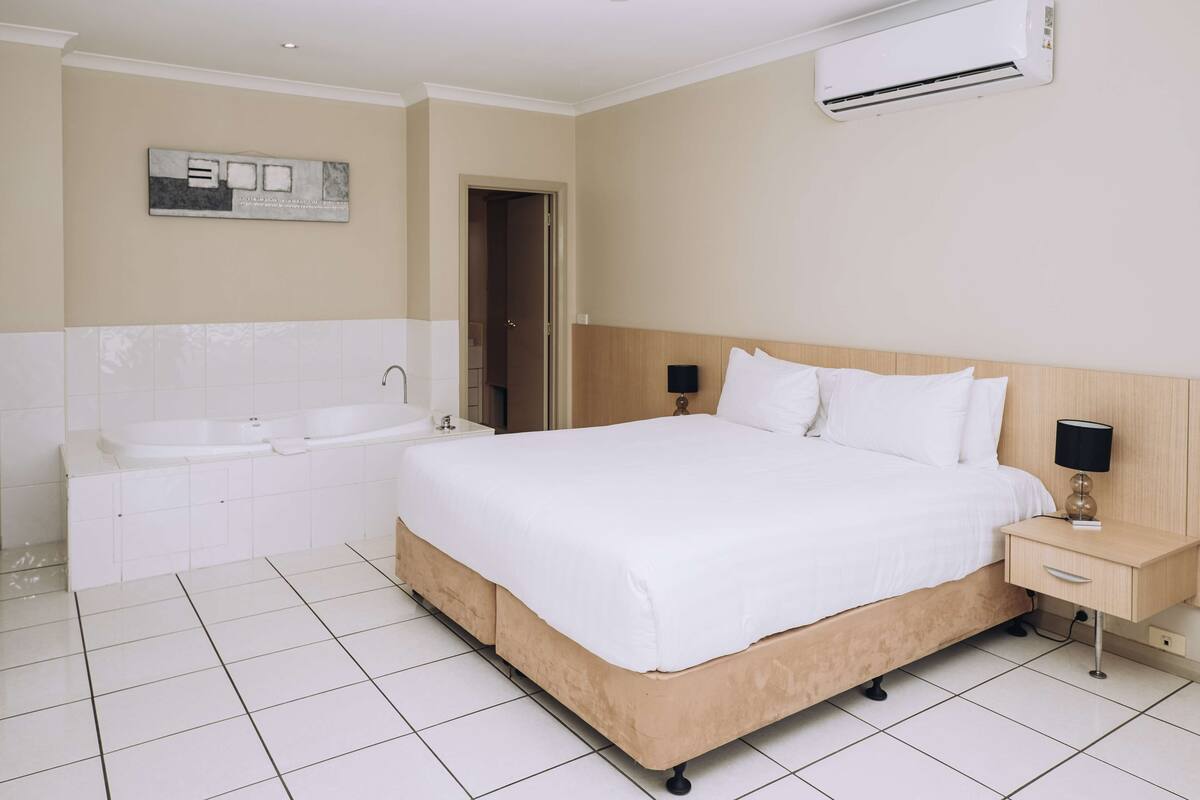 A hotel room with a large bed, a separate bathtub, a bedside table with a lamp, and an air conditioning unit on the wall.