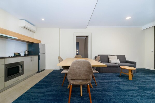 A modern kitchen with a built-in oven and microwave, a dining area with a wooden table and chairs, and a grey sofa with a small round table.