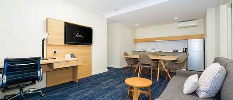 Una habitación de hotel con escritorio, sofá, una mesita redonda, televisor y kitchenette.