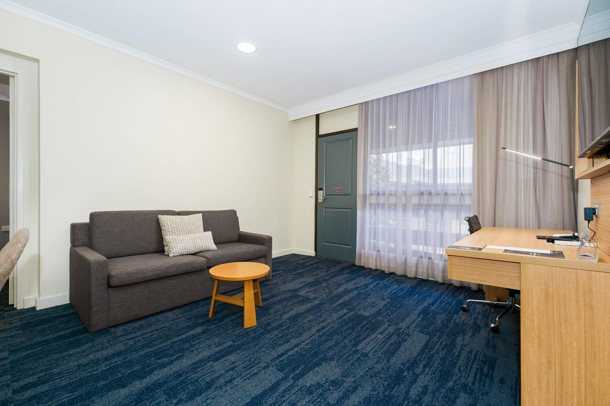 Ein Hotelzimmer mit einer Couch, einem kleinen runden Tisch, einem Schreibtisch mit Stuhl und einem großen Fenster mit Vorhängen.