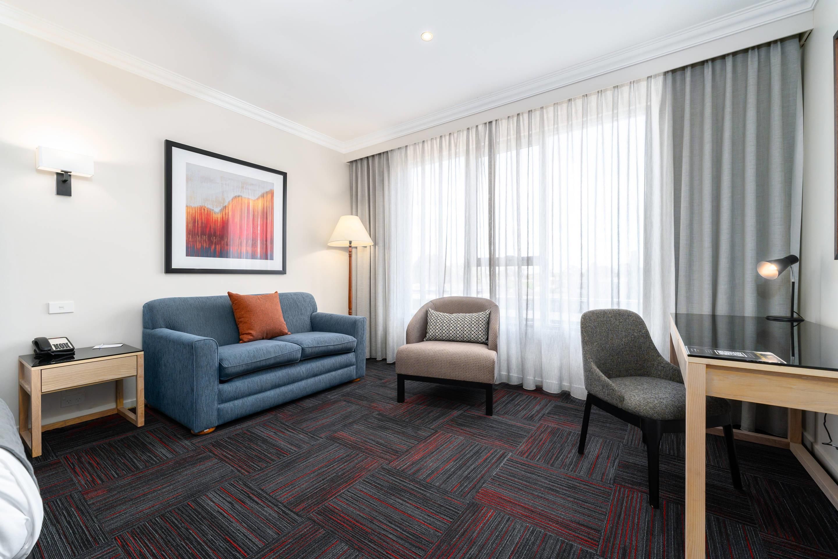 Habitación de hotel con sofá azul, sillón gris, escritorio con lámpara, cuadro abstracto enmarcado y ventana grande con cortinas de gasa.
