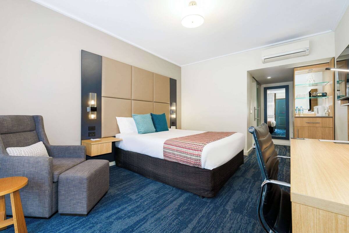 Habitación de hotel con cama, zona de estar con una silla y reposapiés, escritorio y kitchenette.
