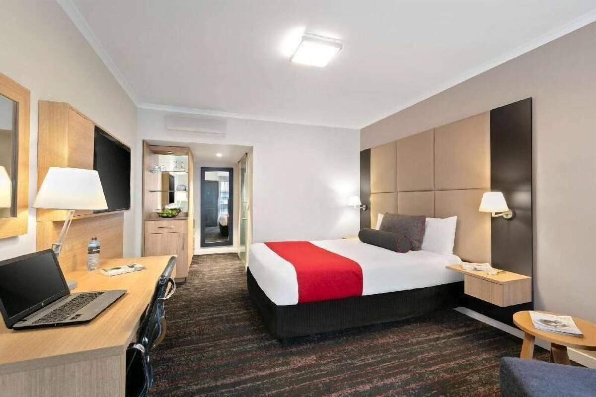 A hotel room with a bed, a desk with a laptop, a bedside table with a lamp, and a small table with a magazine.