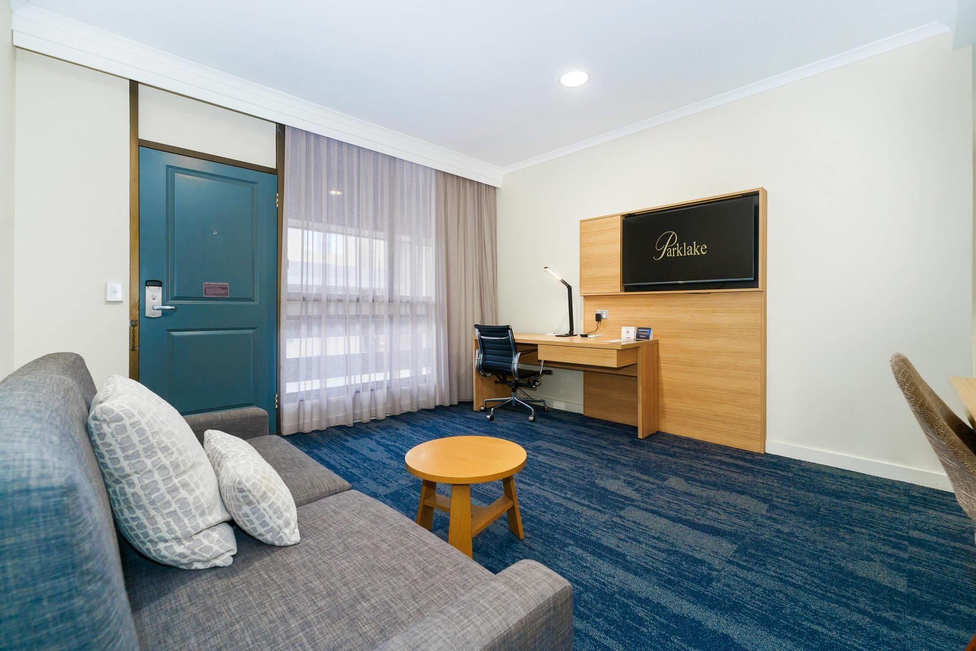 A hotel room with a grey sofa, a small round table, a desk with a chair, a TV, and a door with a curtain.