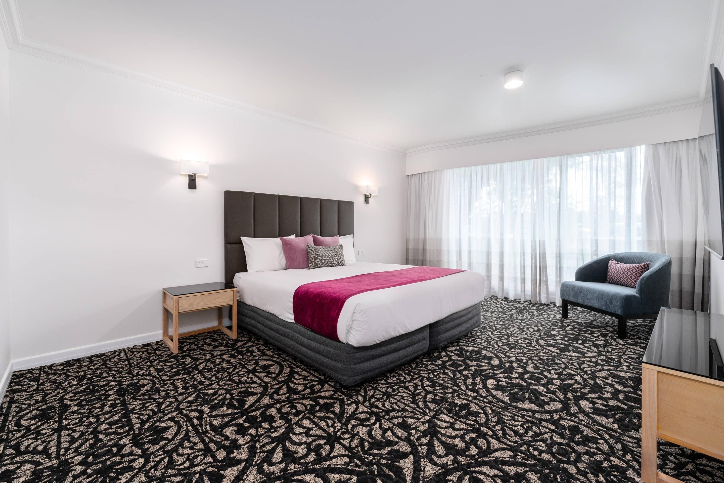 Una habitación de hotel con una cama grande, una mesita de noche, una silla y una ventana con cortinas.