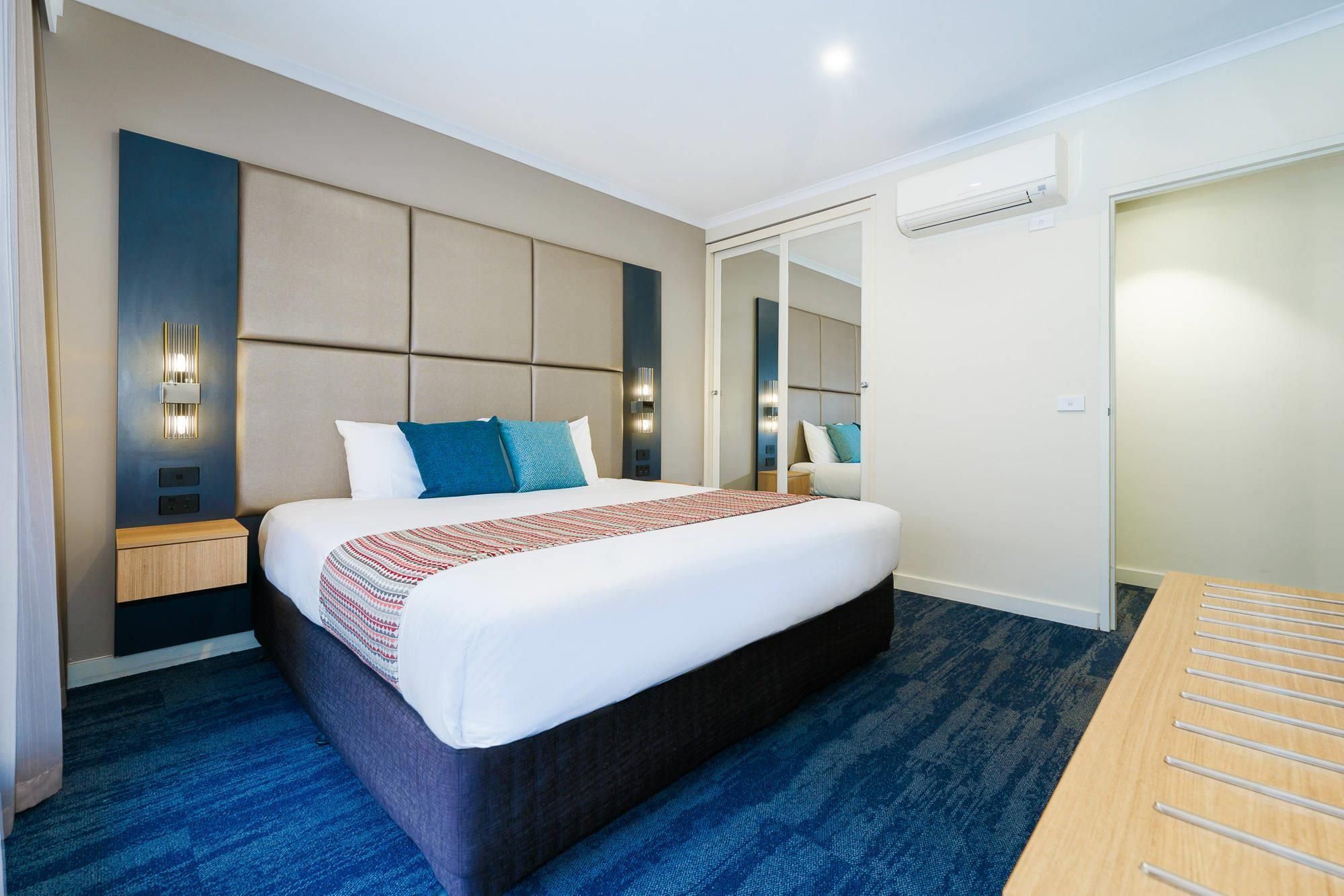A modern hotel room with a large bed, a bedside table, a mirror, and an air conditioning unit.