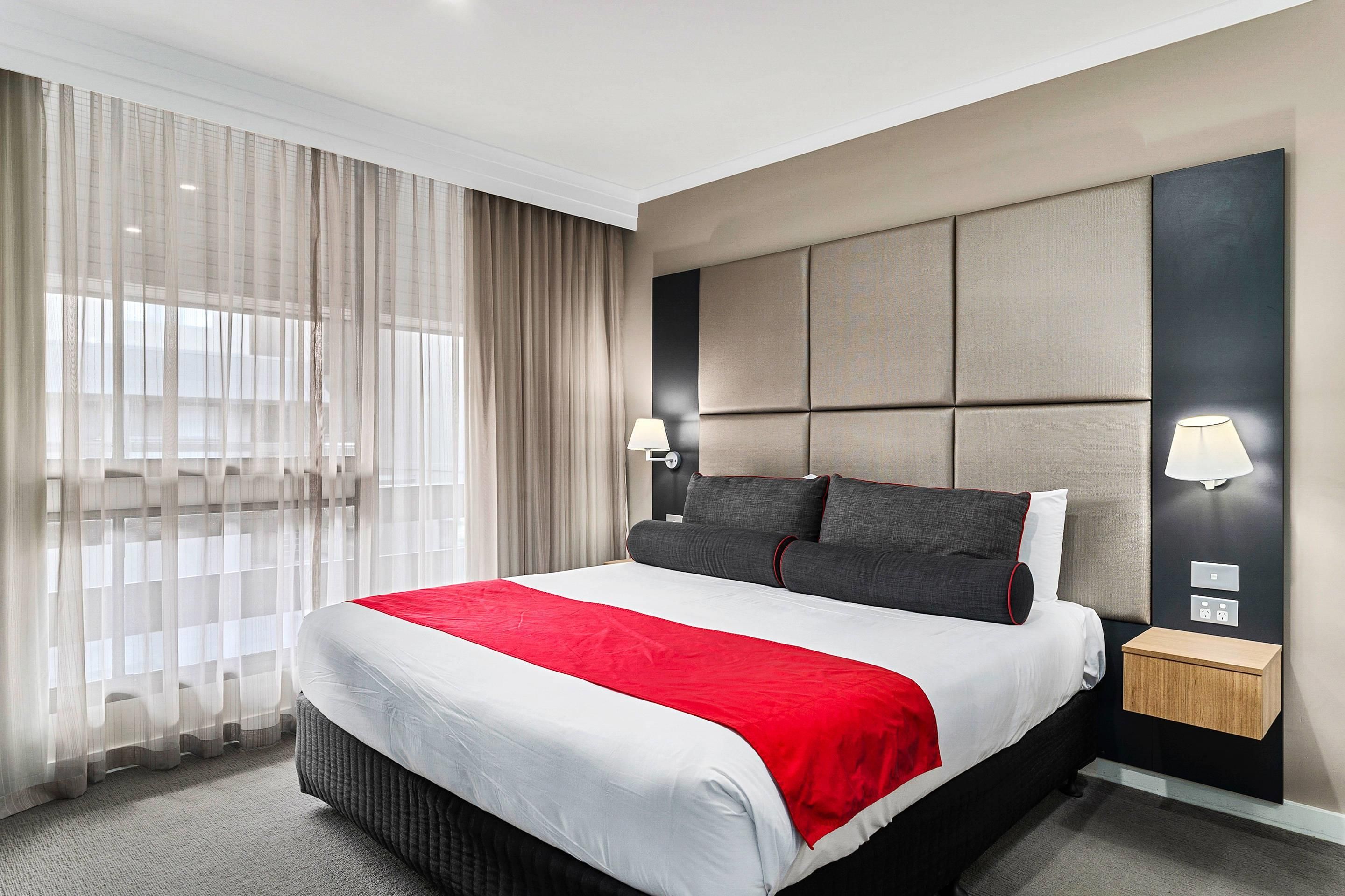 Una habitación de hotel con una cama grande, cabecera con paneles, dos lámparas de noche, mesitas de noche y cortinas traslúcidas.