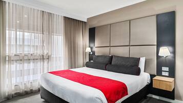Una habitación de hotel con una cama grande, cabecera con paneles, dos lámparas de noche, mesitas de noche y cortinas traslúcidas.