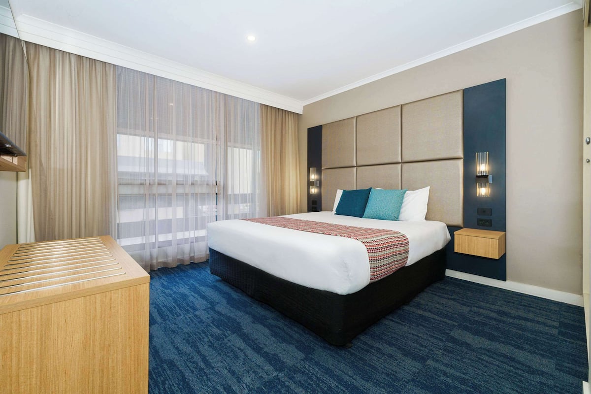 Una habitación de hotel con una cama grande, una mesita de madera, una lámpara de pared y una ventana con cortinas traslúcidas.