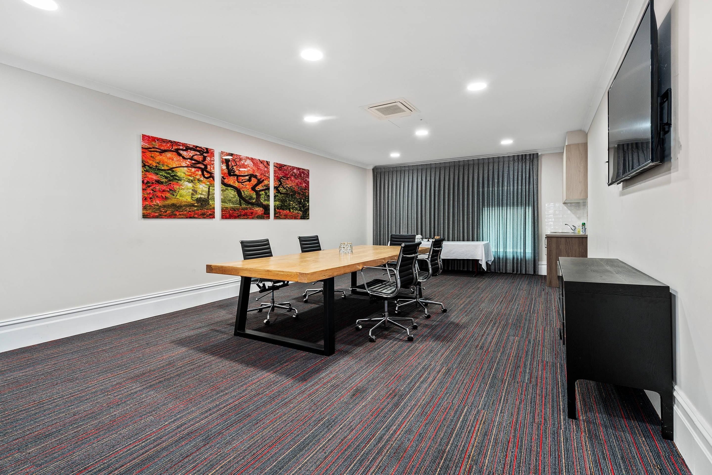 A conference room with a long table, four chairs, a television, and two paintings on the wall.