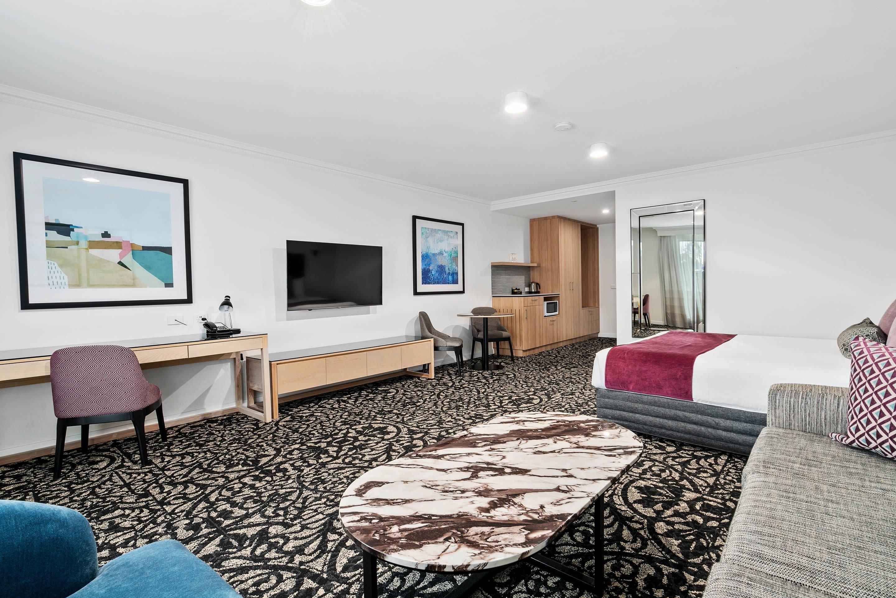 A hotel room with a bed, a desk, a TV, a kitchen area, and a large mirror.