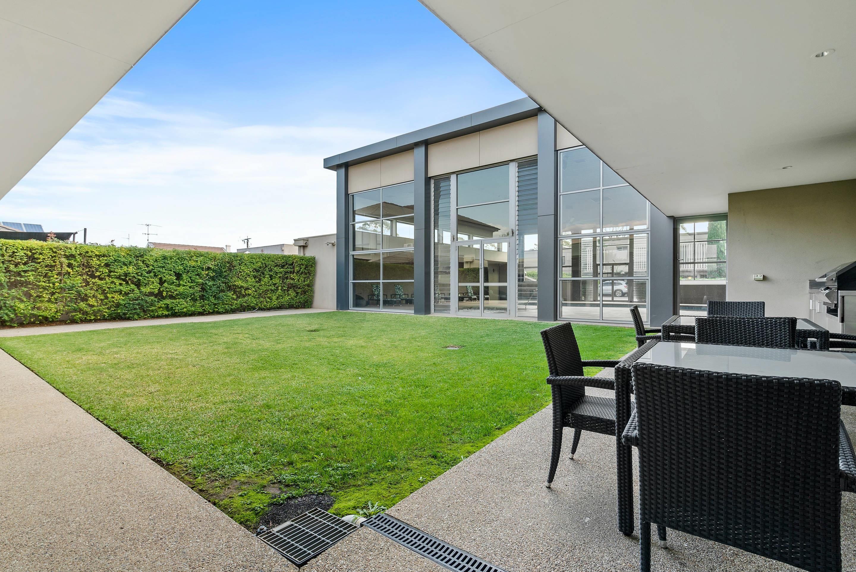 A modern outdoor patio with a grassy lawn, a seating area with wicker furniture, and a glass-enclosed building with large windows.