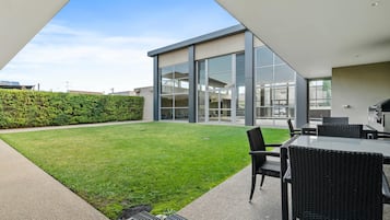 A modern outdoor patio with a grassy lawn, a seating area with wicker furniture, and a glass-enclosed building with large windows.