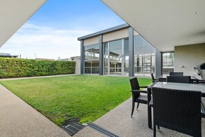 A modern outdoor patio with a grassy lawn, a seating area with wicker furniture, and a glass-enclosed building with large windows.