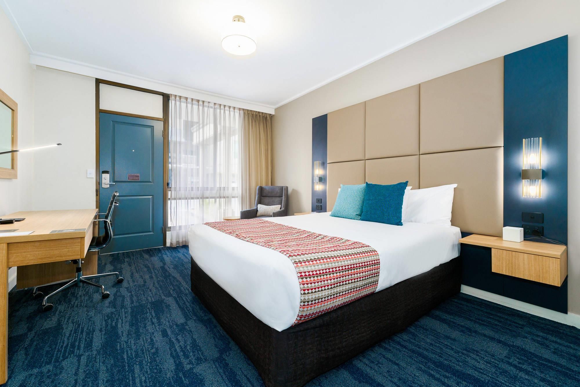 Habitación de hotel con cama, escritorio, silla, mesita de noche y lámpara fijada a la pared.