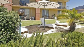 A hotel with a curved red facade, a covered outdoor seating area with white chairs and a large umbrella, and a well-maintained garden with various plants.