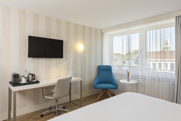 Habitación de hotel con cama, escritorio con silla, televisor, lámpara, mesita de noche con un florero y ventana con cortinas.