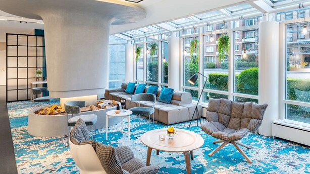 A modern lounge area with a large sofa, armchairs, and a coffee table. There is a fire pit with logs and a small table with a wine glass. The room has large windows with a view of buildings and greenery outside. - NH Hamburg Mitte (Hamburg)
