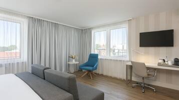 Habitación de hotel con una cama grande, un sofá gris, un sillón azul, un escritorio con silla, un televisor de pantalla plana y ventanas amplias con cortinas.