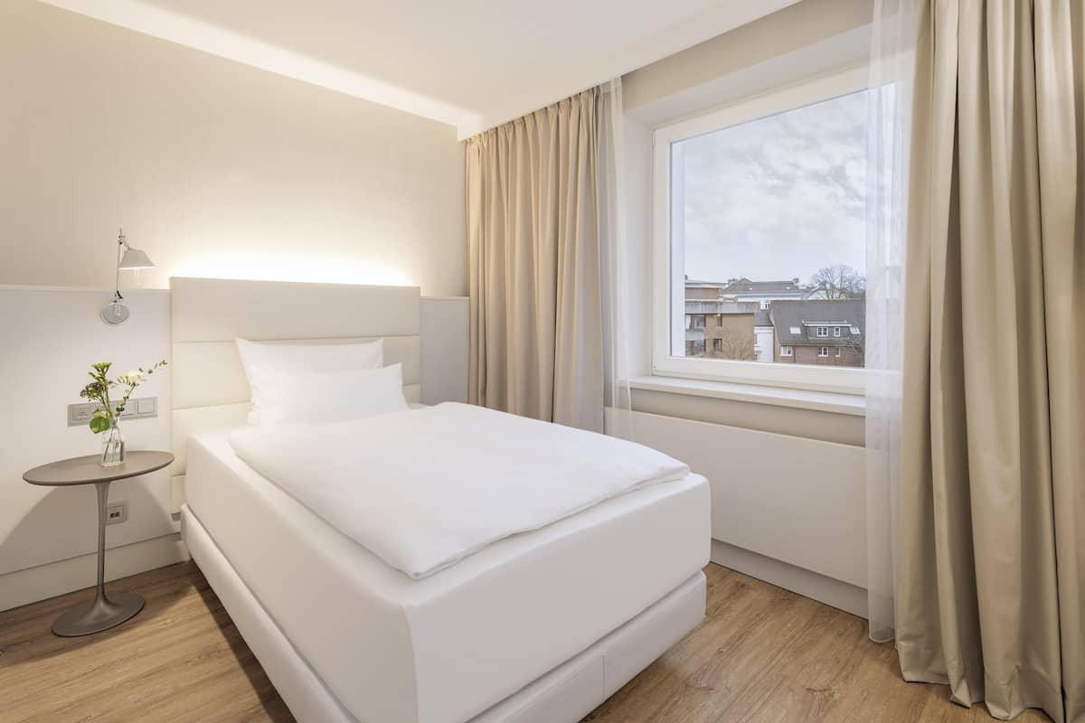 A hotel room with a large bed, a bedside table with a lamp, a window with curtains, and a view of buildings outside.