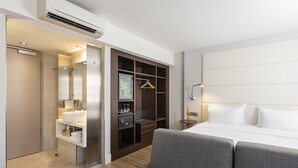 A modern hotel room with a large bed, a grey sofa, a walk-in closet, a bathroom with a sink and mirror, and an air conditioning unit. - NH Hamburg Mitte (Hamburg)