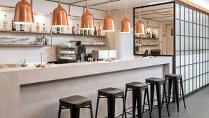 A modern bar area with copper pendant lights, bar stools, and shelves stocked with bottles and glasses. - NH Hamburg Mitte (Hamburg)