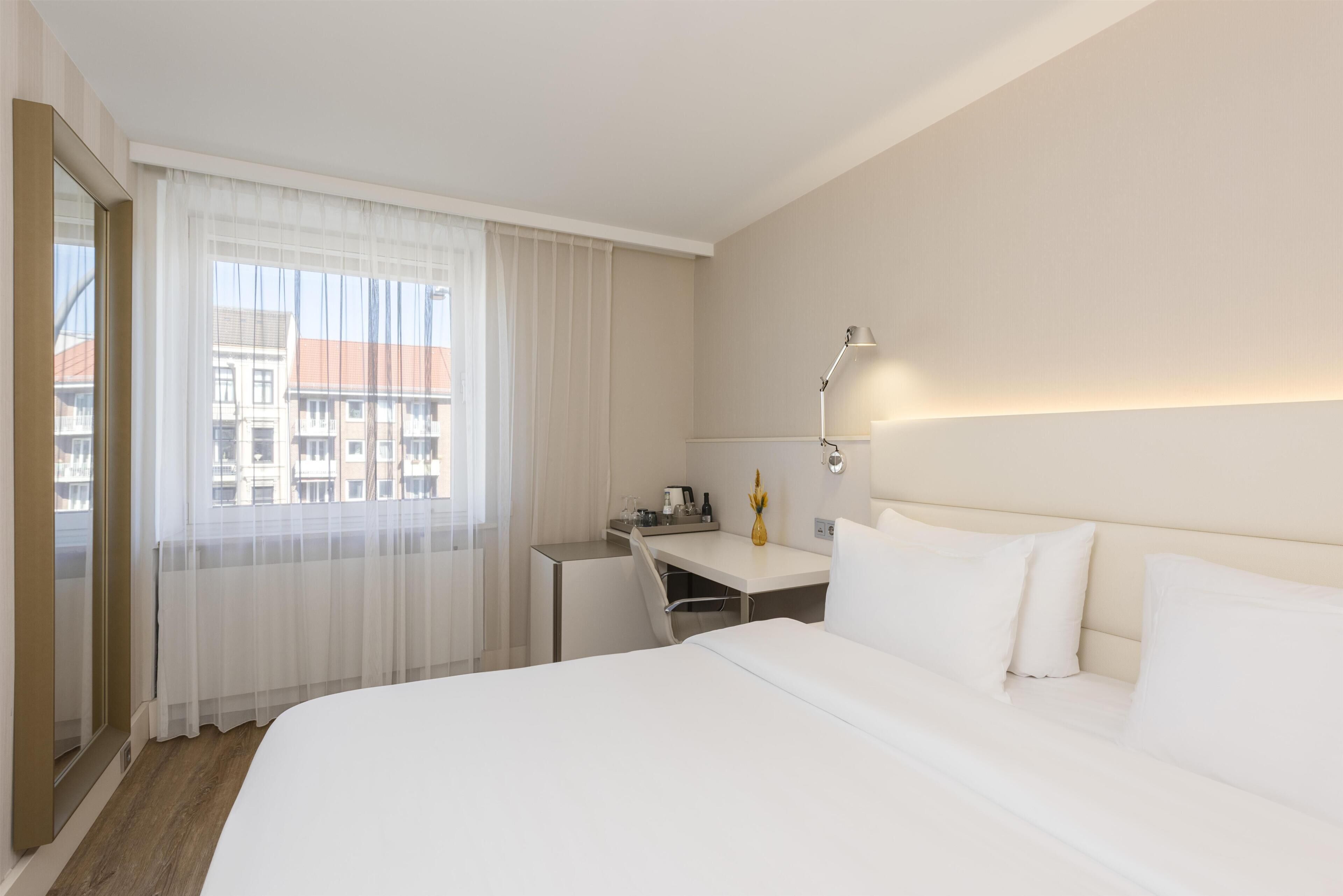 A hotel room with a bed, a desk, a chair, a lamp, and a window with curtains.