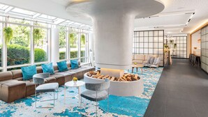 A modern lobby with a large central column, seating area, and a view of greenery outside. - NH Hamburg Mitte (Hamburg)
