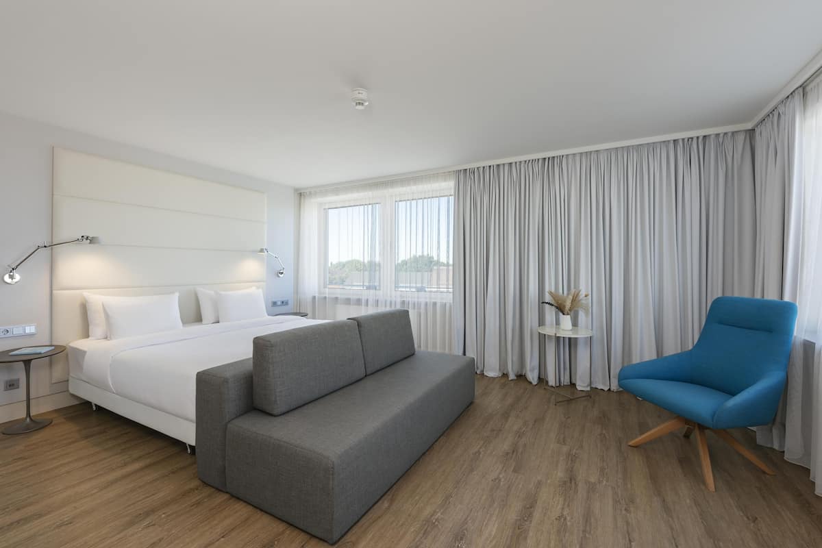 A modern hotel room with a bed, a grey sofa, a blue armchair, a small side table, and a window with curtains.