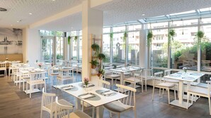 A spacious dining area with white tables and chairs, large windows, and hanging plants. - NH Hamburg Mitte (Hamburg)
