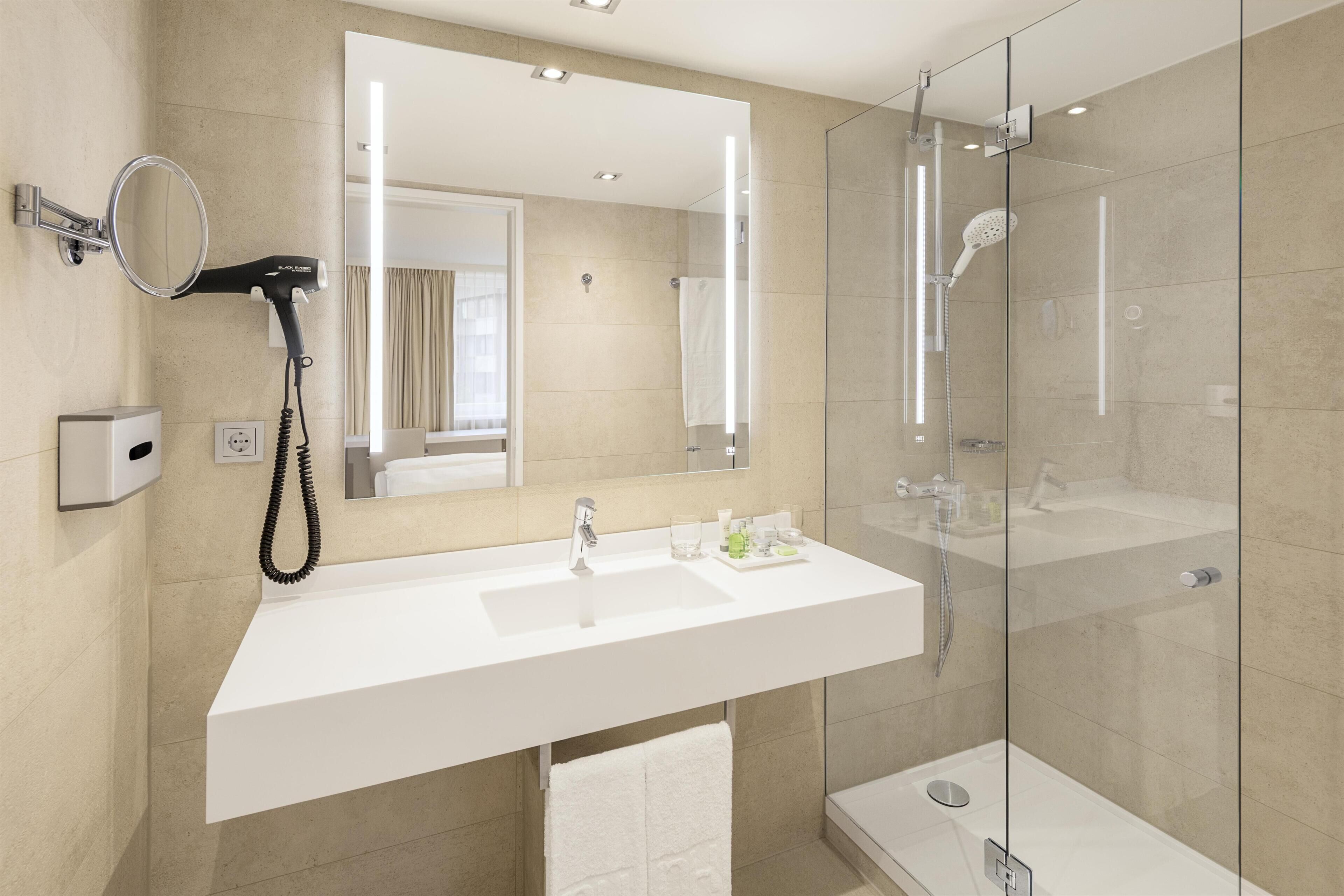 Un baño moderno con un espejo grande, un secador de pelo, un lavamanos y una ducha con mampara de vidrio.