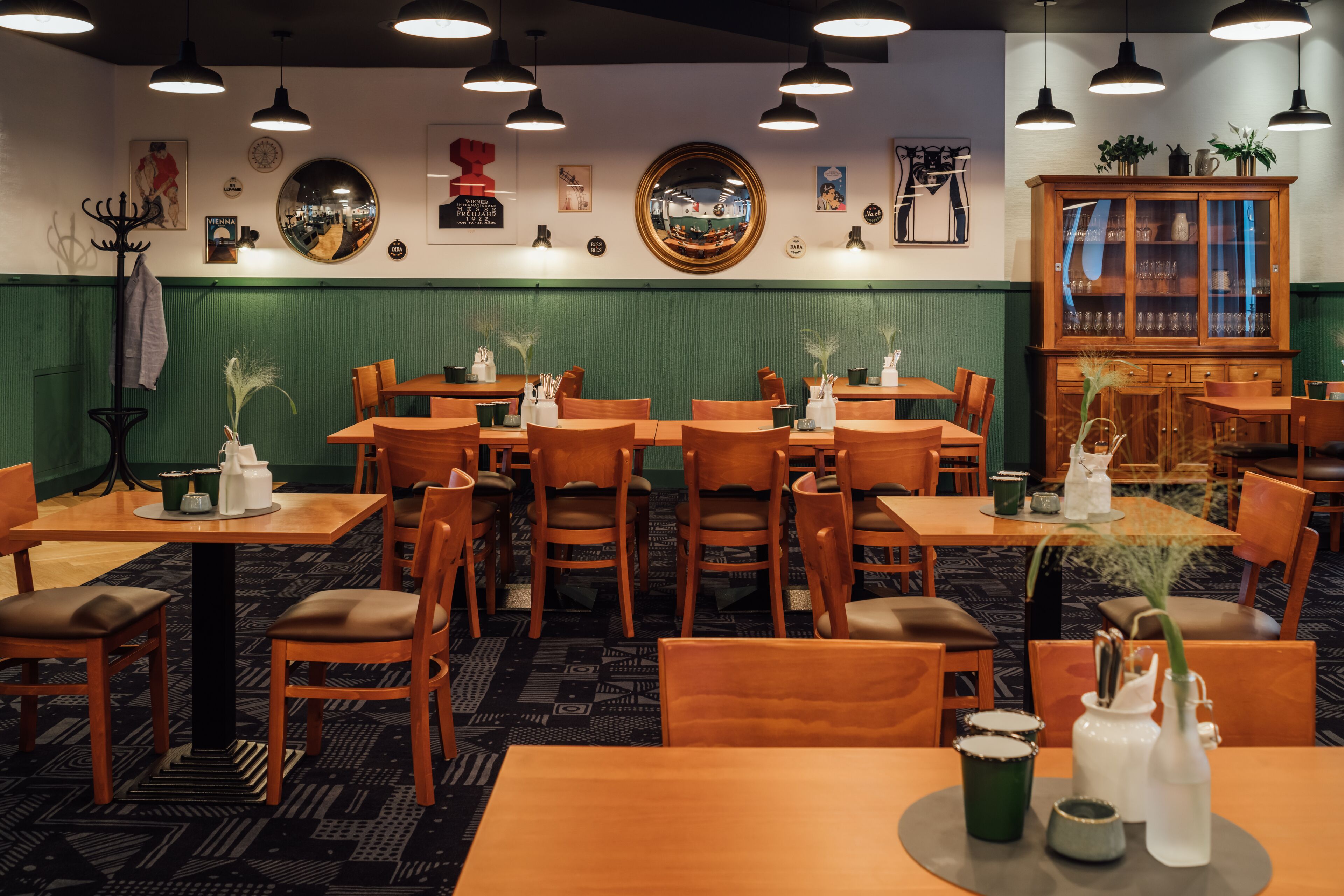 A restaurant interior with wooden tables and chairs, hanging pendant lights, and a green paneled wall.