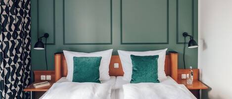 Cama de casal com roupa de cama branca e almofadas verdes, duas mesas de cabeceira com luminárias e cortina com padrão geométrico.