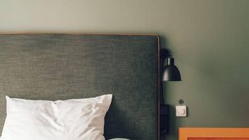 Ein ordentlich bezogenes Bett mit weißen Leinen, einem grünen Kissen und einem hölzernen Kopfteil. Ein kleiner Nachttisch mit einer Lampe, einem Buch und einer Tasse. An der Wand befinden sich drei große, dunkle, abstrakte Wandbilder.