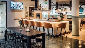 Eine moderne Bar mit einer langen Theke, mehreren Barhocker und einem Bildschirm, der 'VIENNA HOUSE' zeigt.
