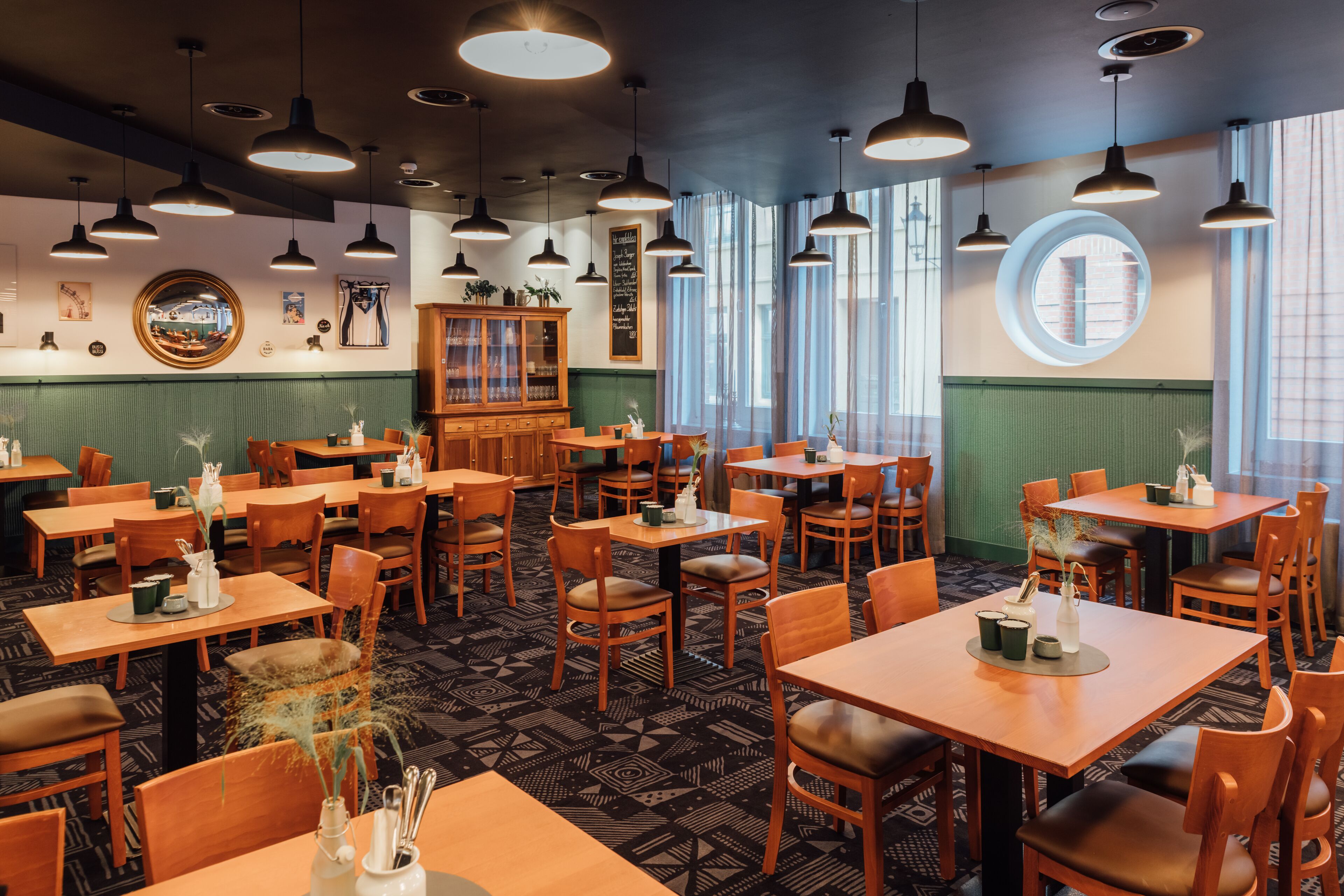 Un restaurant avec des tables et des chaises en bois, des luminaires suspendus et un mur végétal agrémenté d’un miroir et d’œuvres d’art.