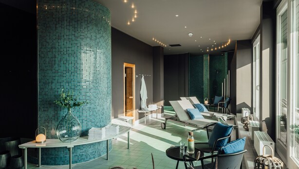 Ein moderner Wellnessbereich mit einer gewölbten, blauen Wand aus Fliesen, Liegestühlen und einem Tisch mit einer Flasche und Gläsern.
